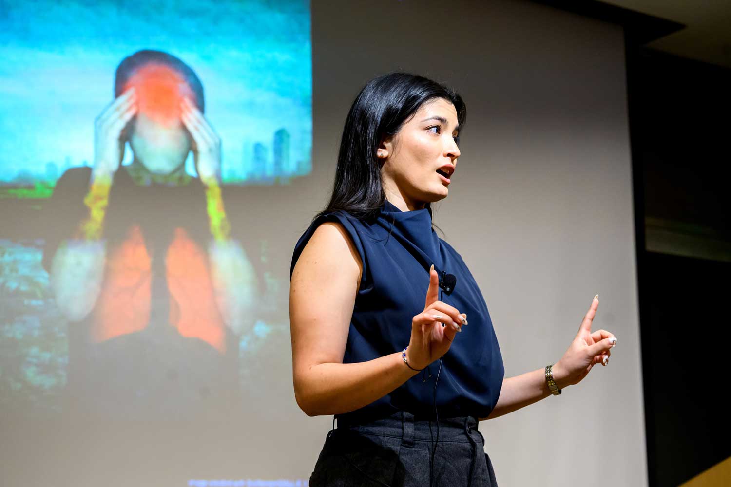 2026 Grad Slam finalist Christine Boutros delivers her talk onstage in front of a projection screen showing an illustration of a person with a headache.