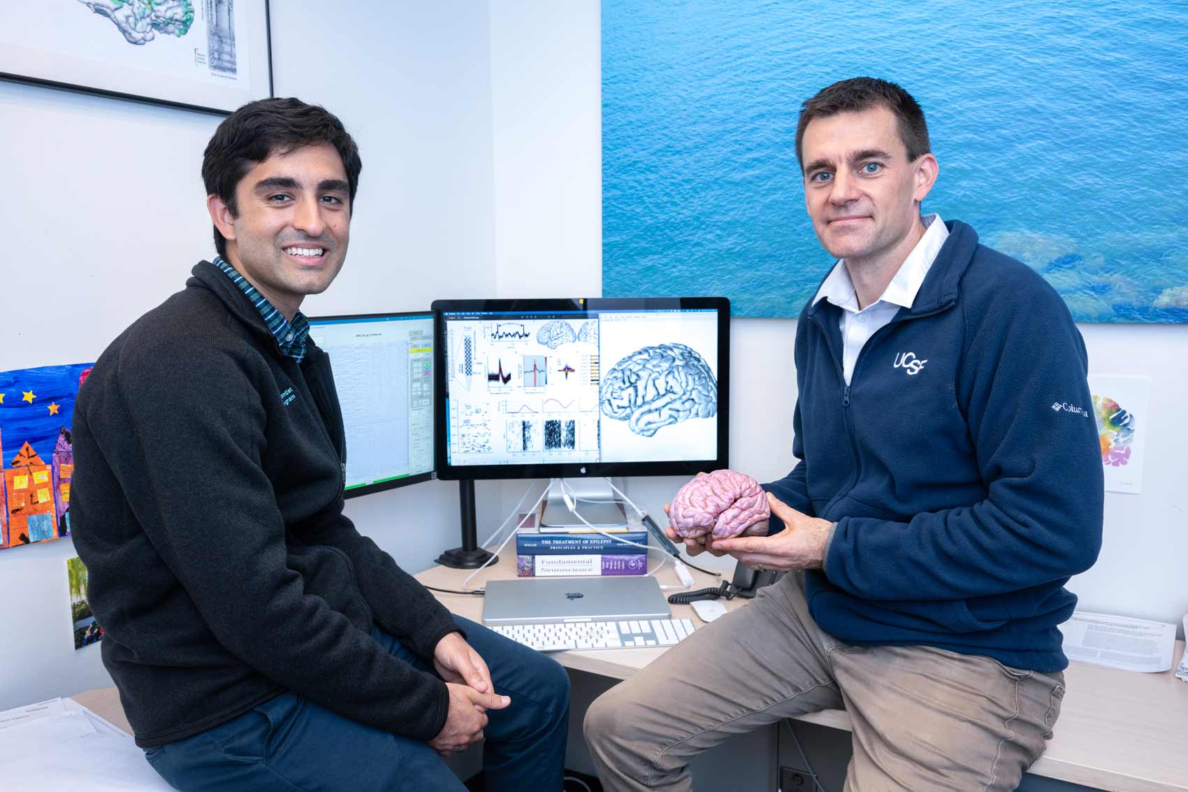 Researchers Jon Kleen and Alex Silva sit in front of a computer showing a 3D model of the brain, along with graphical data used in their research. Jon Kleen holds a model of a brain.