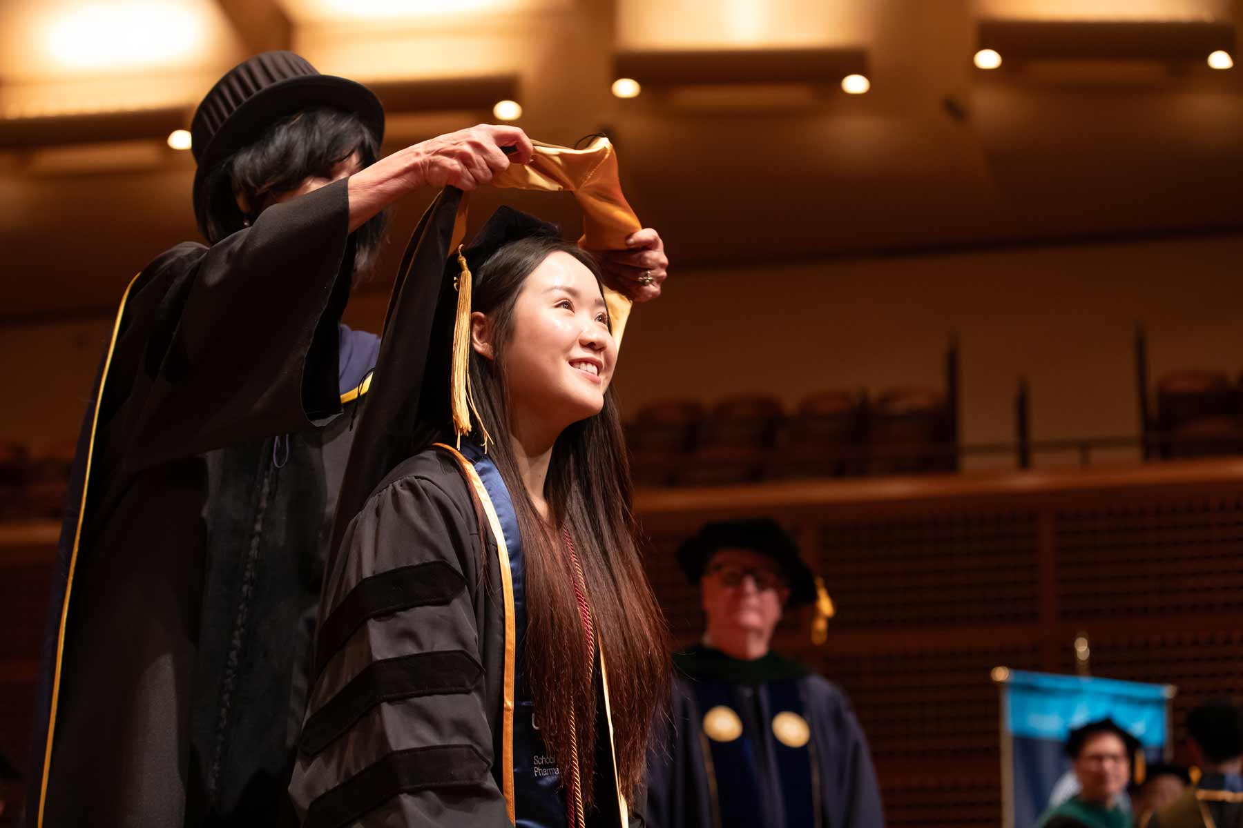 A female school of pharmacy graduate is ceremonially hooded on stage.