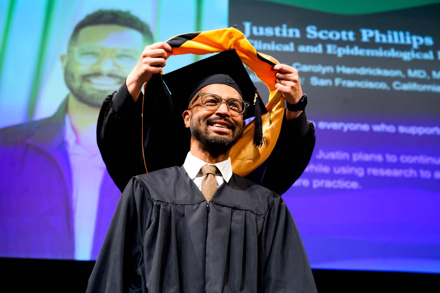 A graduate smiles as he is hooded on stage at commencement.