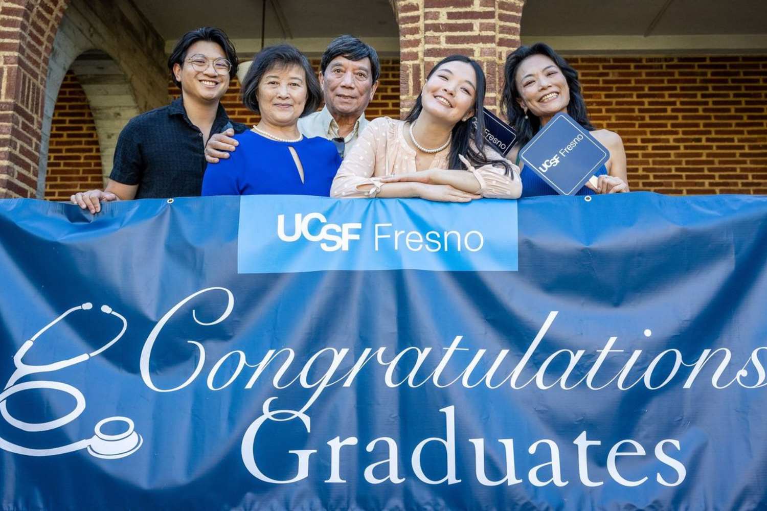 A graduate and their family smiling and posing together in front of a blue UCSF Fresno "Congratulations Graduates" banner