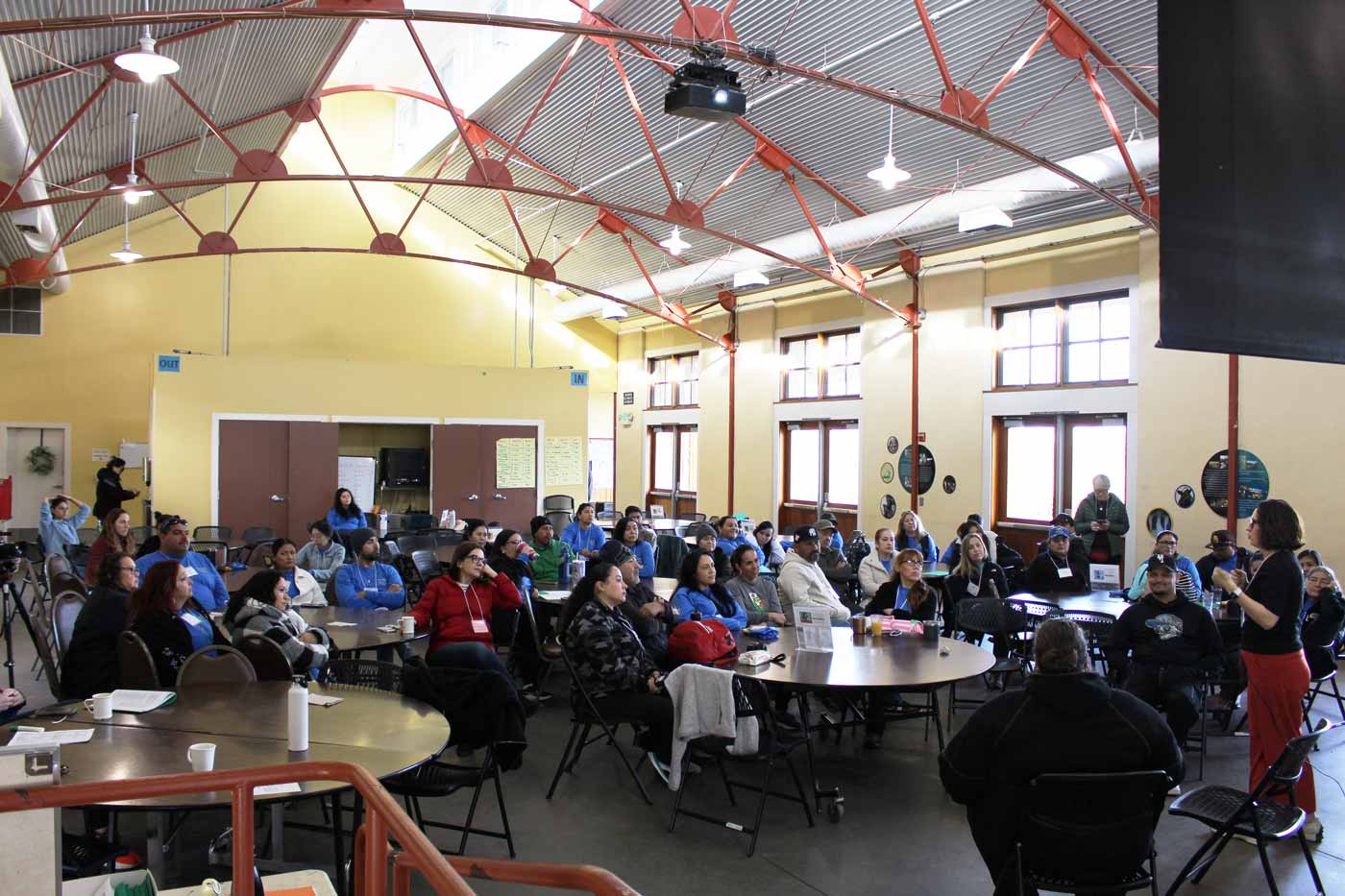 Attendees sit in an assembly room at a campground space while a speaker presents.