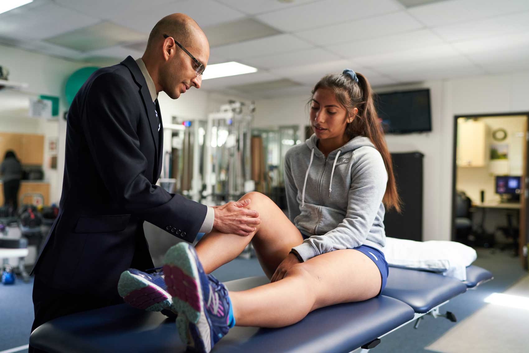 Orthopedic surgeon Nirav Panya examines a surgical scar on a patient's knee inside a physical therapy room.
