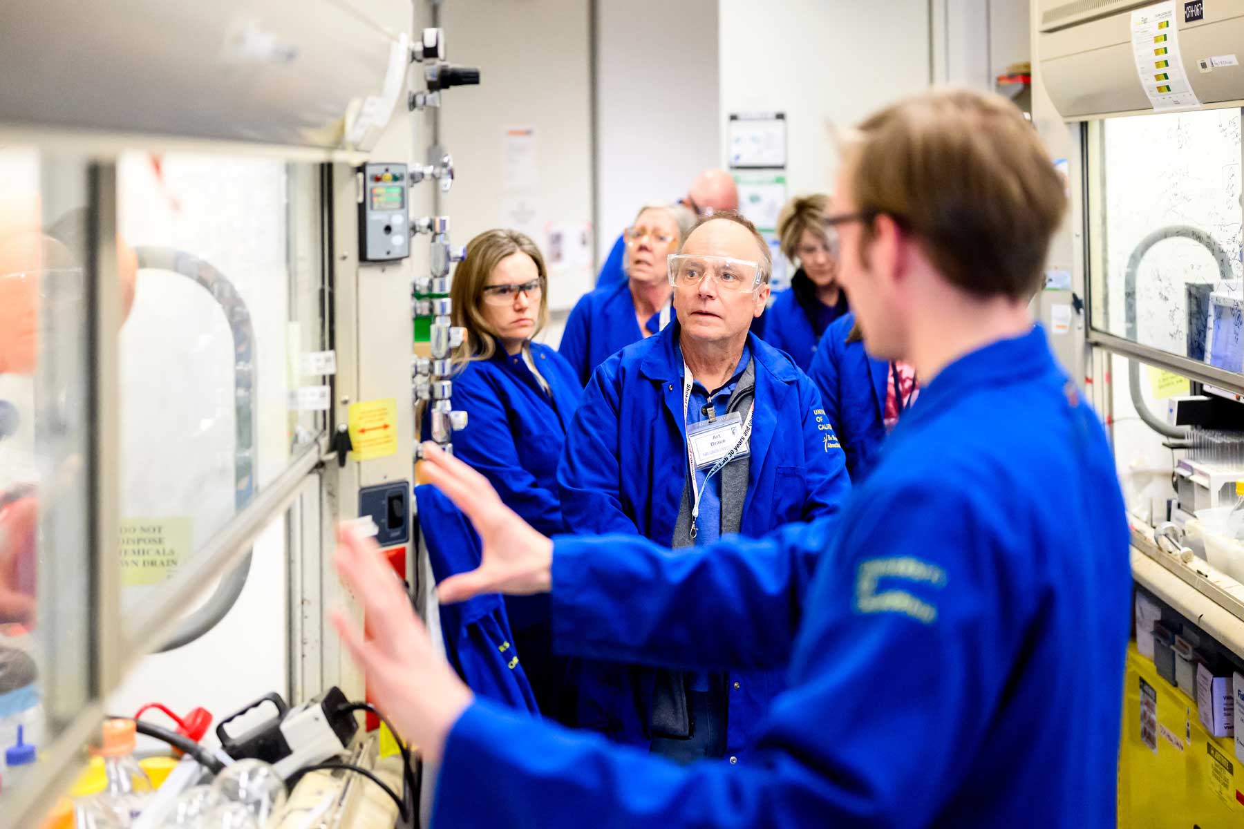 A group wearing blue lab coats and protective goggles tour a lab while a postdoc shows them his work.