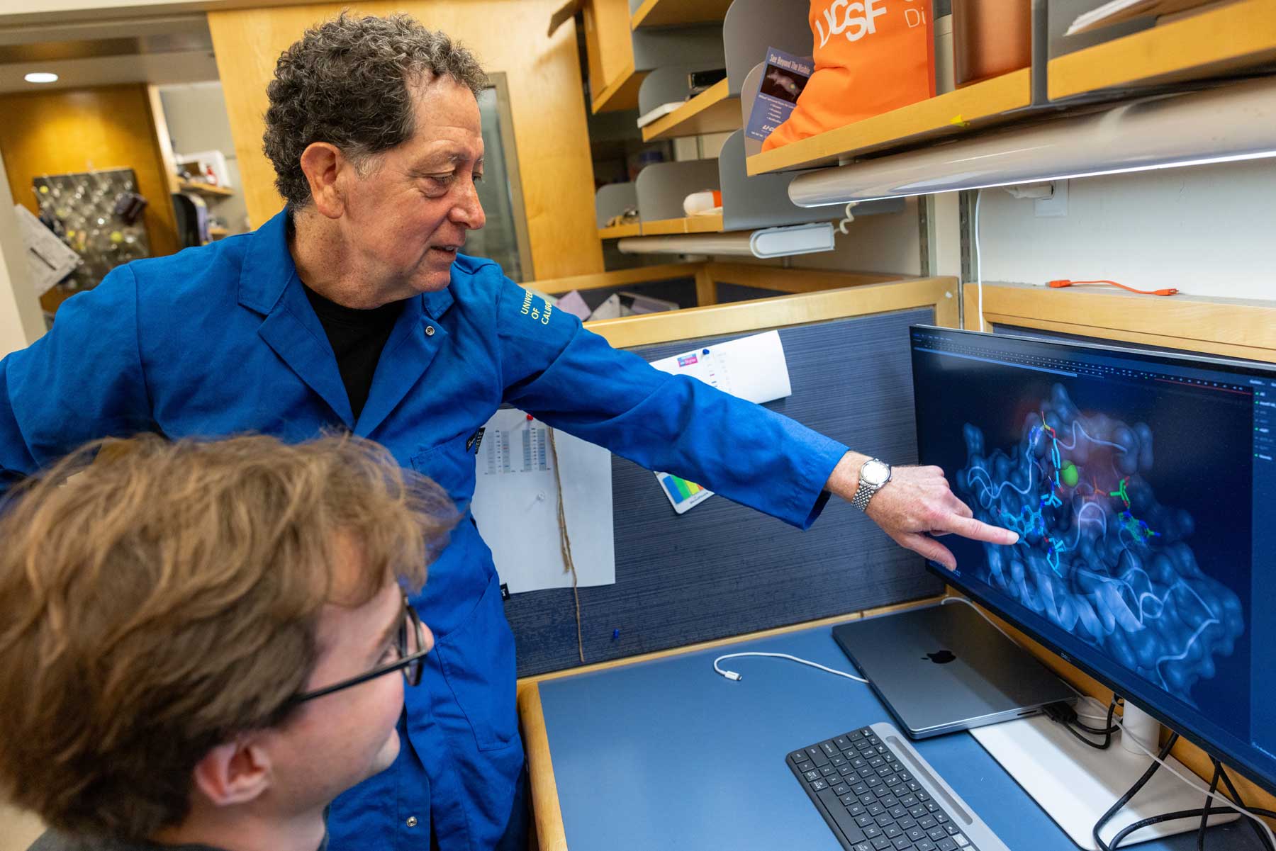 Kevan Shokat points to a 3D molecular model of a KRAS Q61H inhibitor on a computer screen as a graduate student looks on.