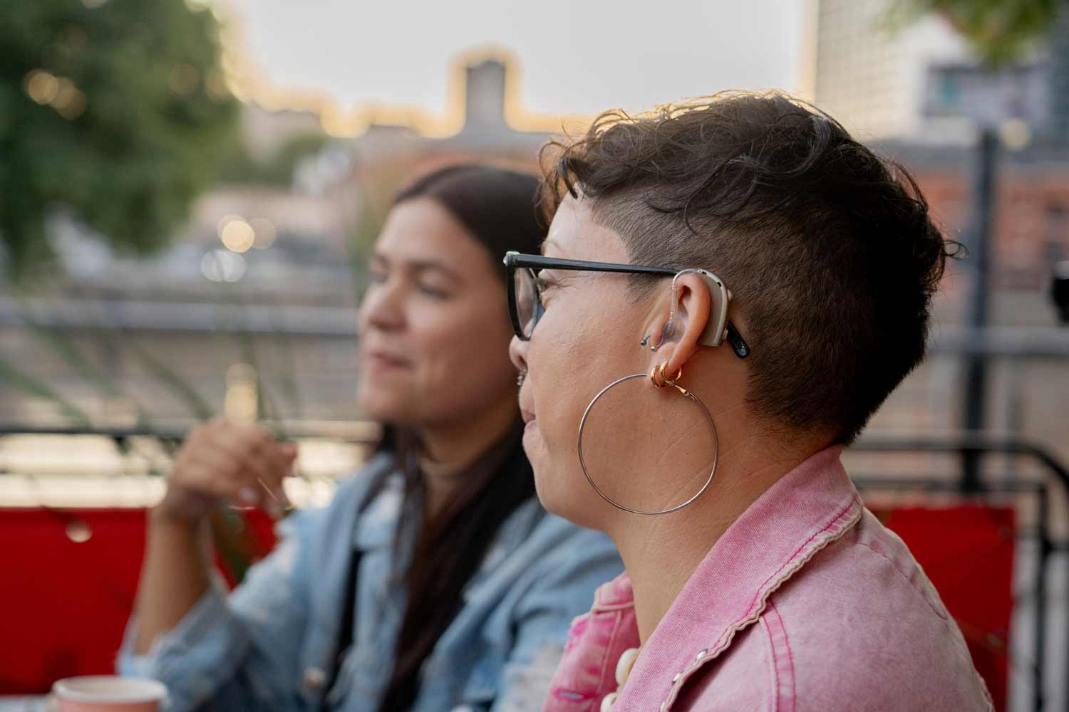 A woman wearing a hearing aid sits outdoors with a companion.