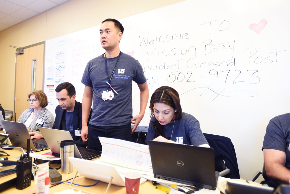 At an Incident Command Post, Chau Vu (standing), director of UCSF Medical Center Emergency Management, watches a GPS screen tracking all the ambulances. The command center also includes (from left to right) M.J. White; Russ Cucina, MD; and Tina Mammone.