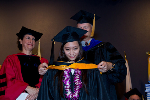 A graduate gets hooded from a faculty member while Dean Elizabeth Watkins looks on.