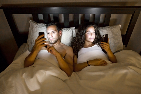 A man and a woman both look at smartphones while in bed