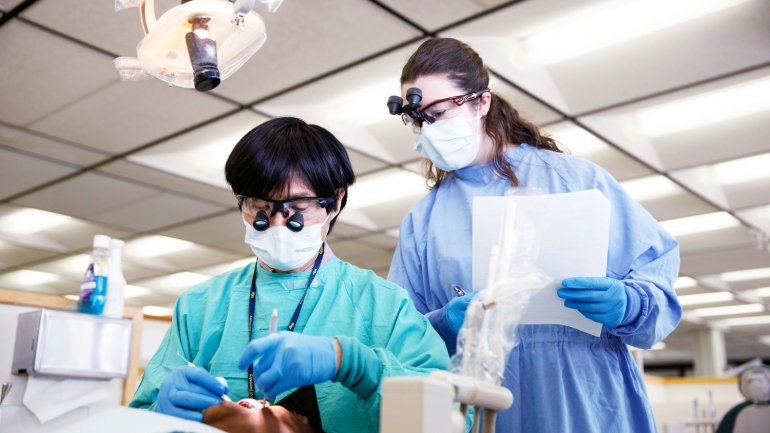 A female dental student stands over a professor to observe him doing a procedure on a patient