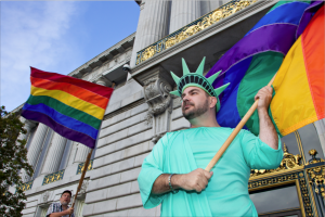 Nikolas Lemos, PhD, dressed as Lady LIberty waiving a rainbow flag outside SF City Hall.