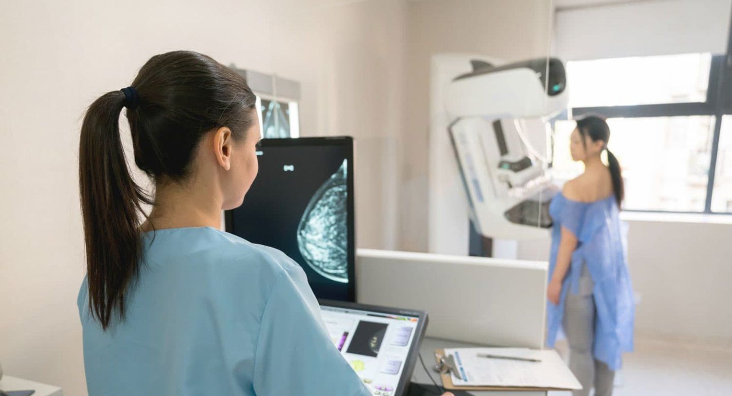 A technician takes a mammogram. The woman getting the mammogram stands at the far end of the room. The scan can be seen on the computer screen.