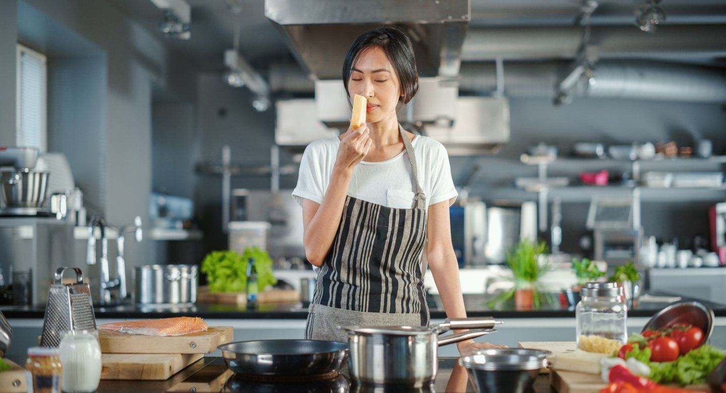 A female chef holds a slice of cheese as she sniffs it. She stands in a large kitchen