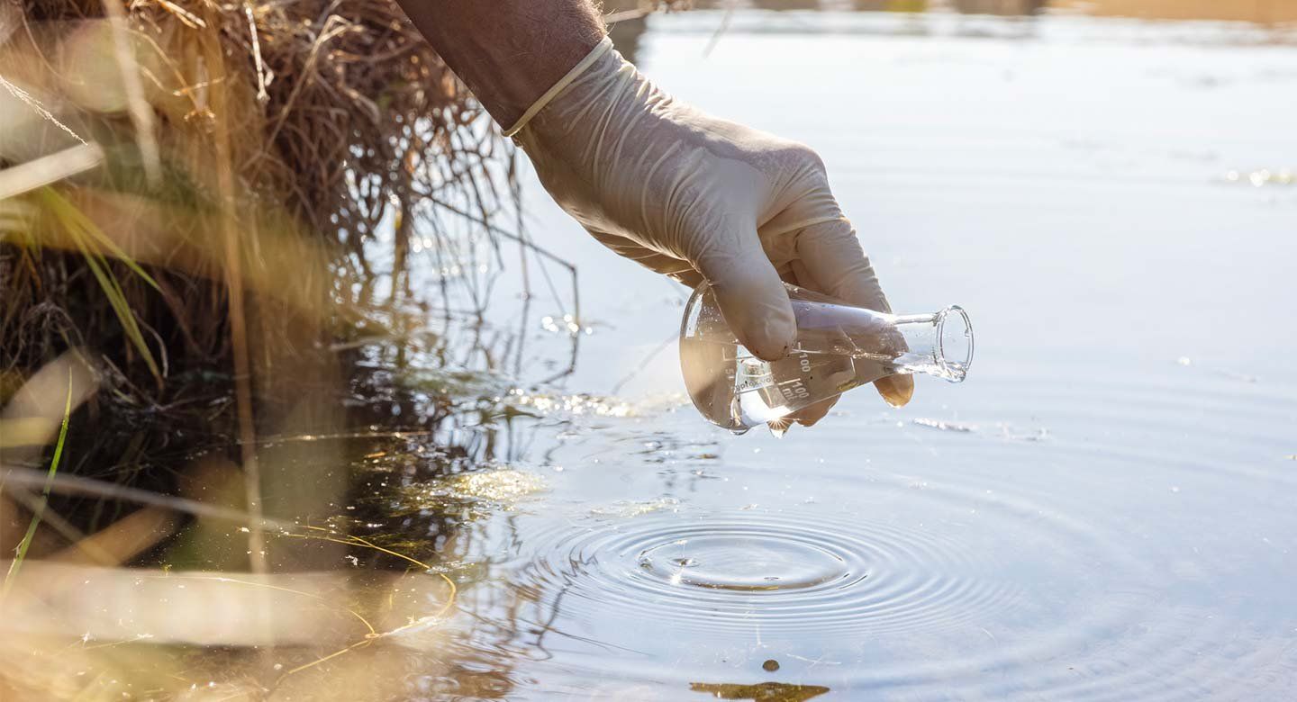 A gloved hand dips a laboratory flask into a lake to collect a water sample for testing.