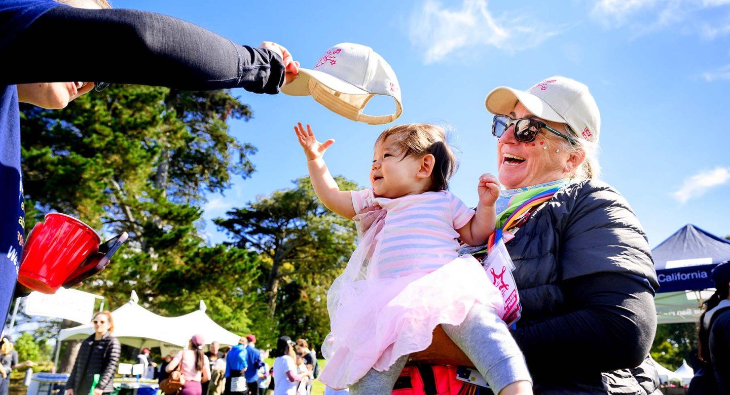 Someone reaches out to put a baseball cap onto a baby girl held by beaming a UCSFer during the event.