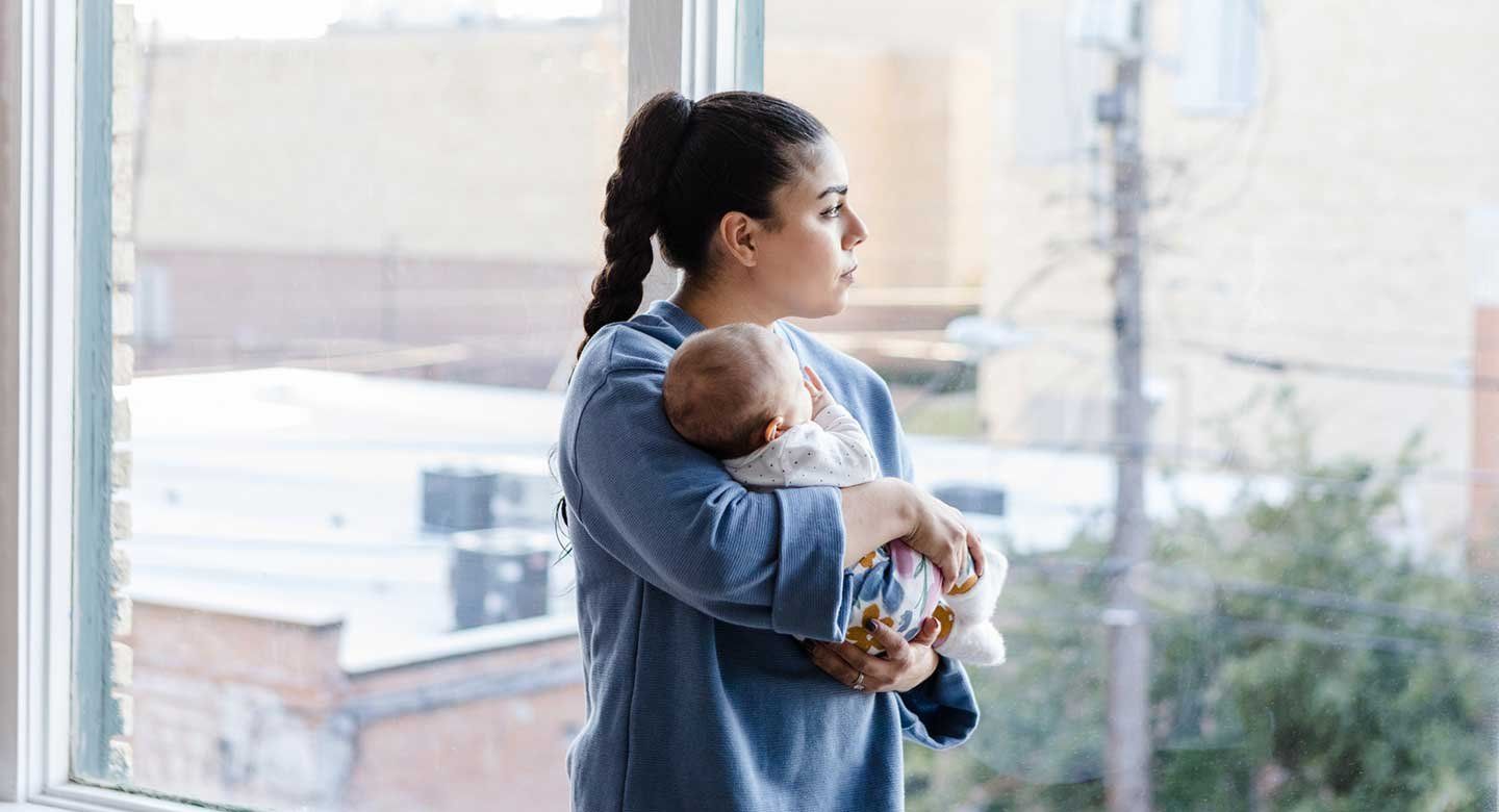 A mother holds her baby and looks contemplatively out of a large, light-filled window
