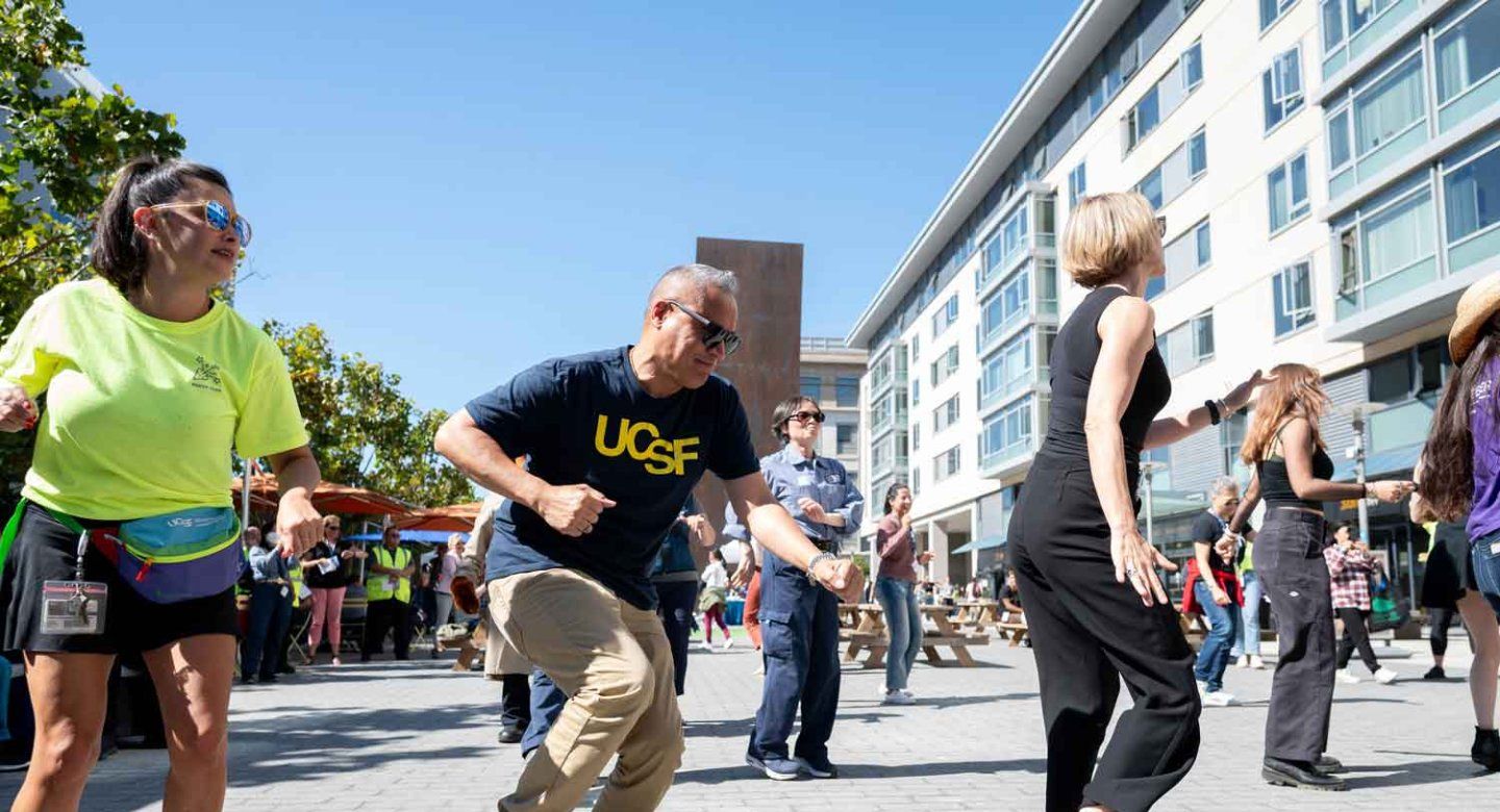 A diverse group of UCSF employees do a line dance outdoors under a bright blue sky.