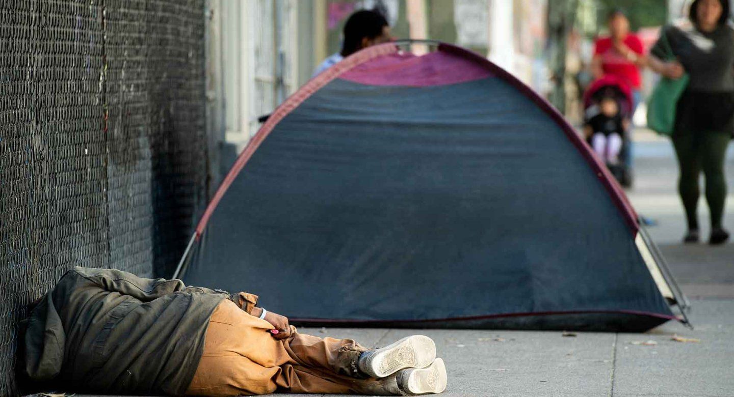 A homless person sleeps on a sidewalk next to a tent.
