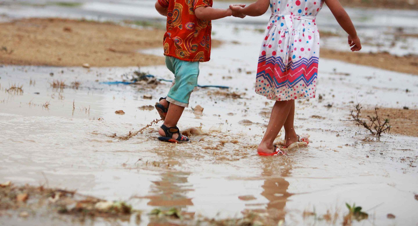 A toddler-aged boy and girl walk together through muddy water after strong rains.