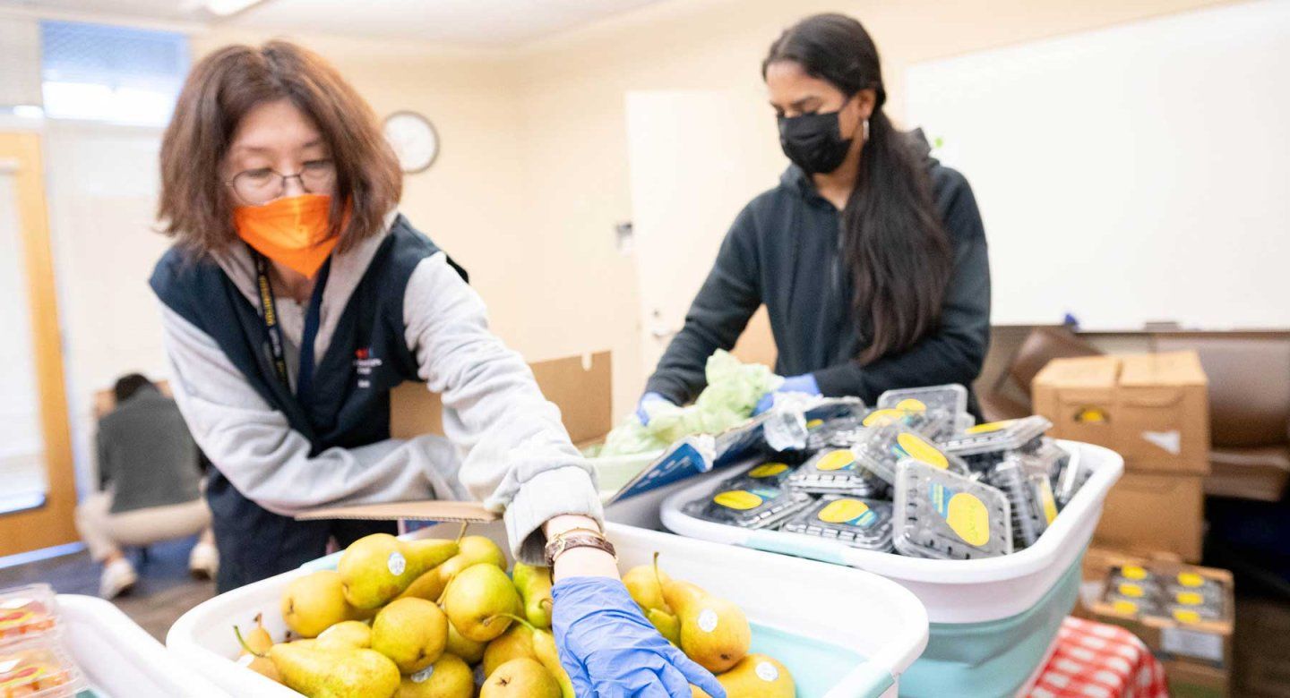 Two volunteers wearing facial masks stand in front of bins filled with pears and blueberries and put some of the fruit into plastic bags.