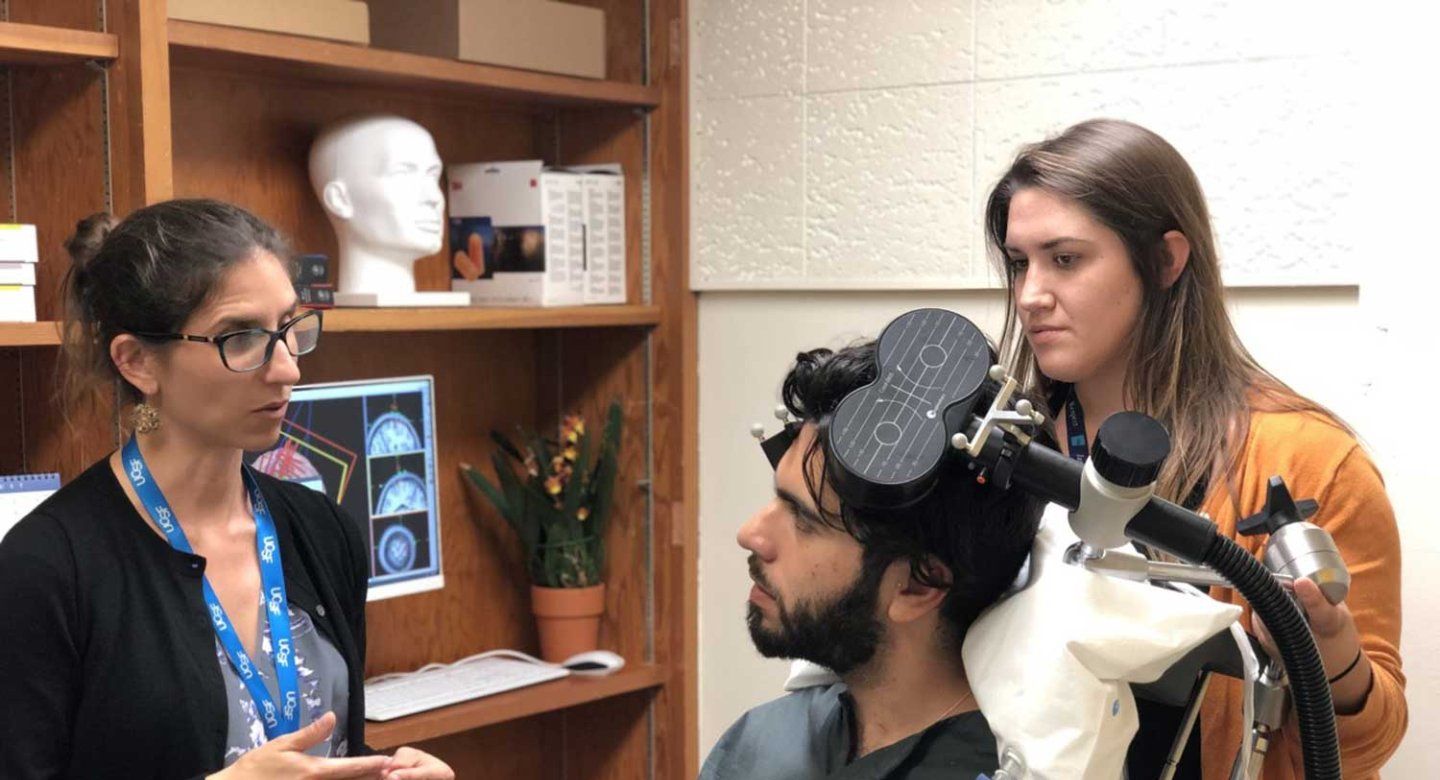 Psychiatrist Katherine Scangos explains the transcranial magnetic simulation (T M S) procedure to a man sitting on an examination chair. Above his head is a device used for the treatment.
