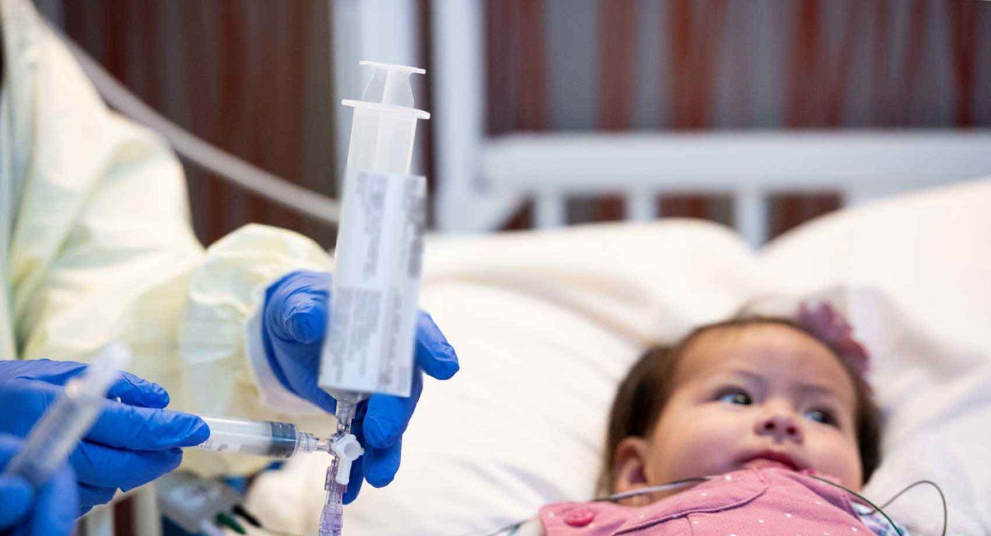 A baby is given medication through an IV by a medical professional wearing protective gear.