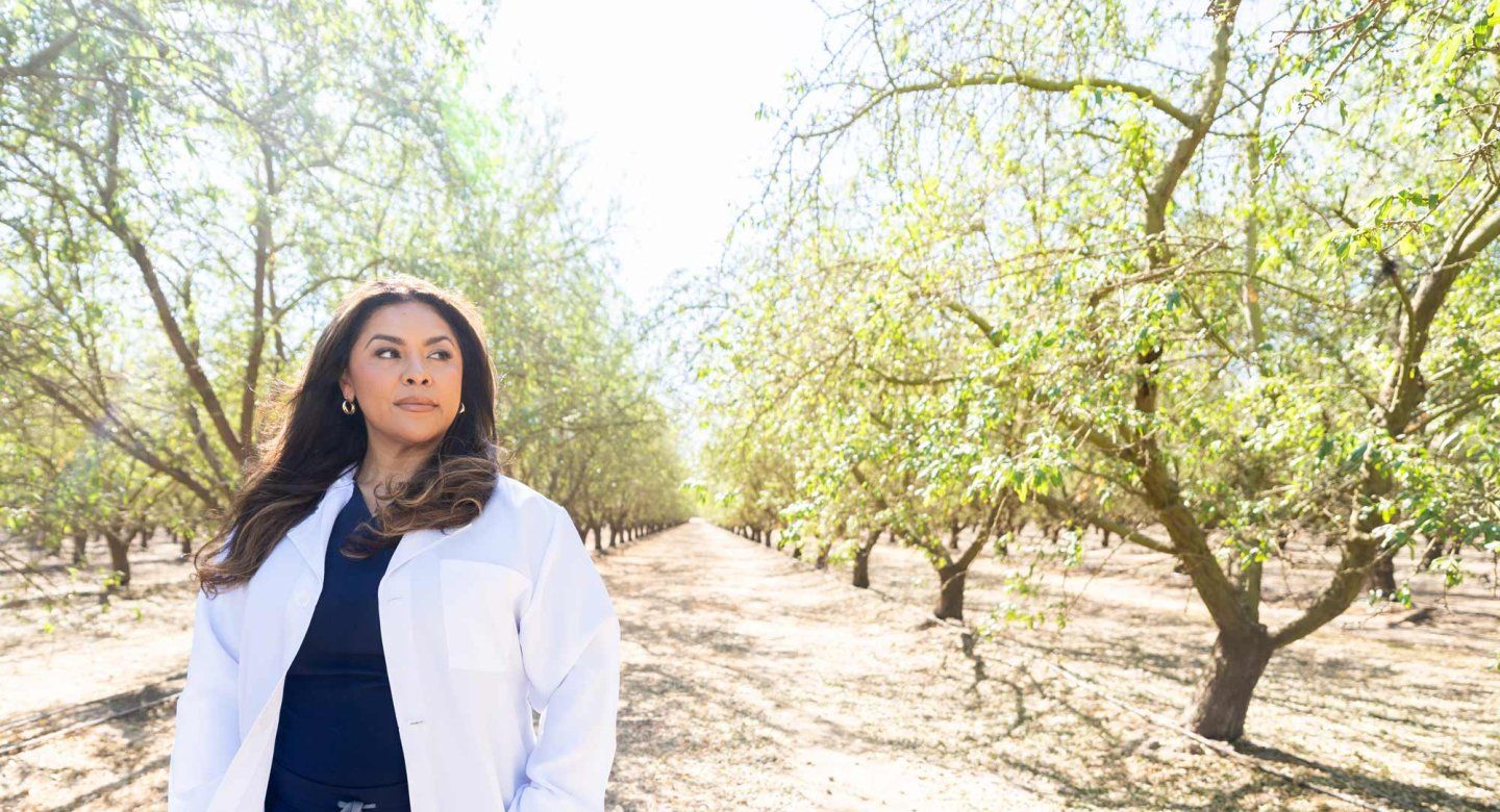 Pharmacy technician Bibiana Arroyo wears a white coat as she walks through an agricultural orchard of trees in Modesto.