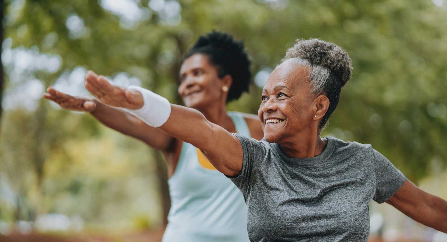 Two Black senior-aged women smile as they do exercise outdoors.
