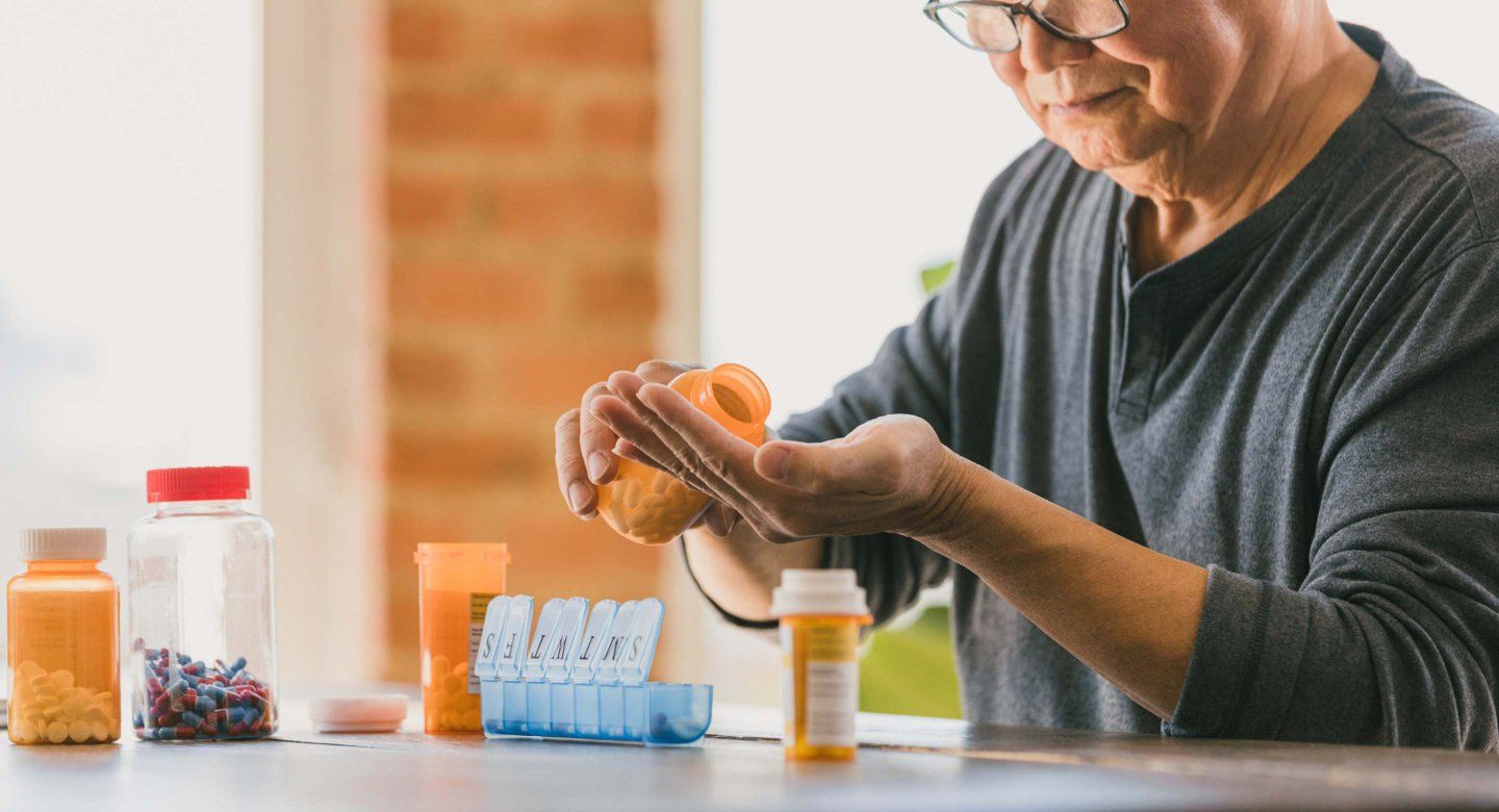 Elderly person with glasses pouring prescription pills from an orange bottle into their hand next to a weekly pill organizer.