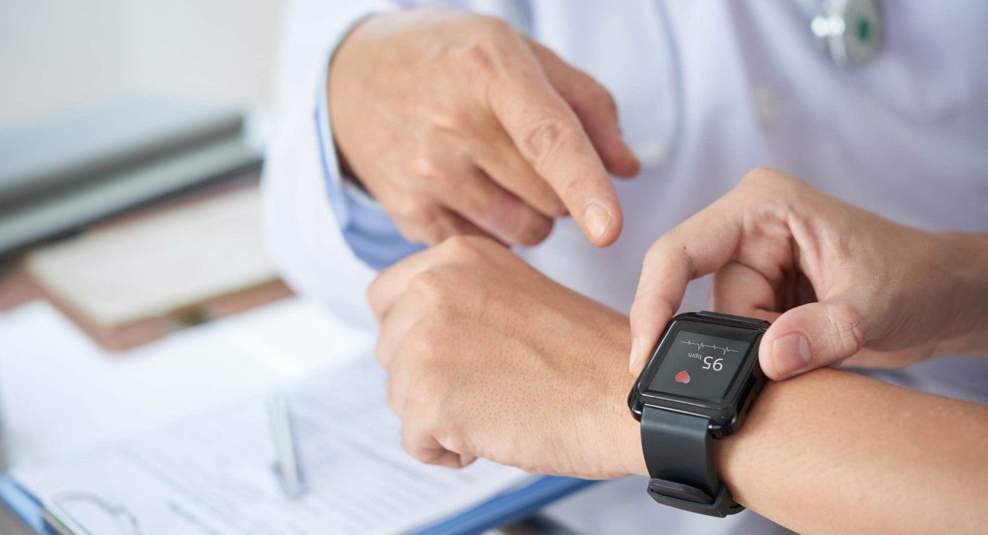 A doctor points to a person's wrist wearing a smartwatch displaying a heart rate of 95 bpm, with paperwork in the background.