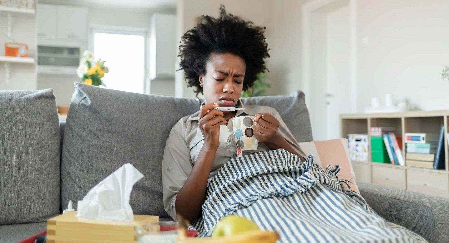 A sick woman takes her temperature on her couch at home.