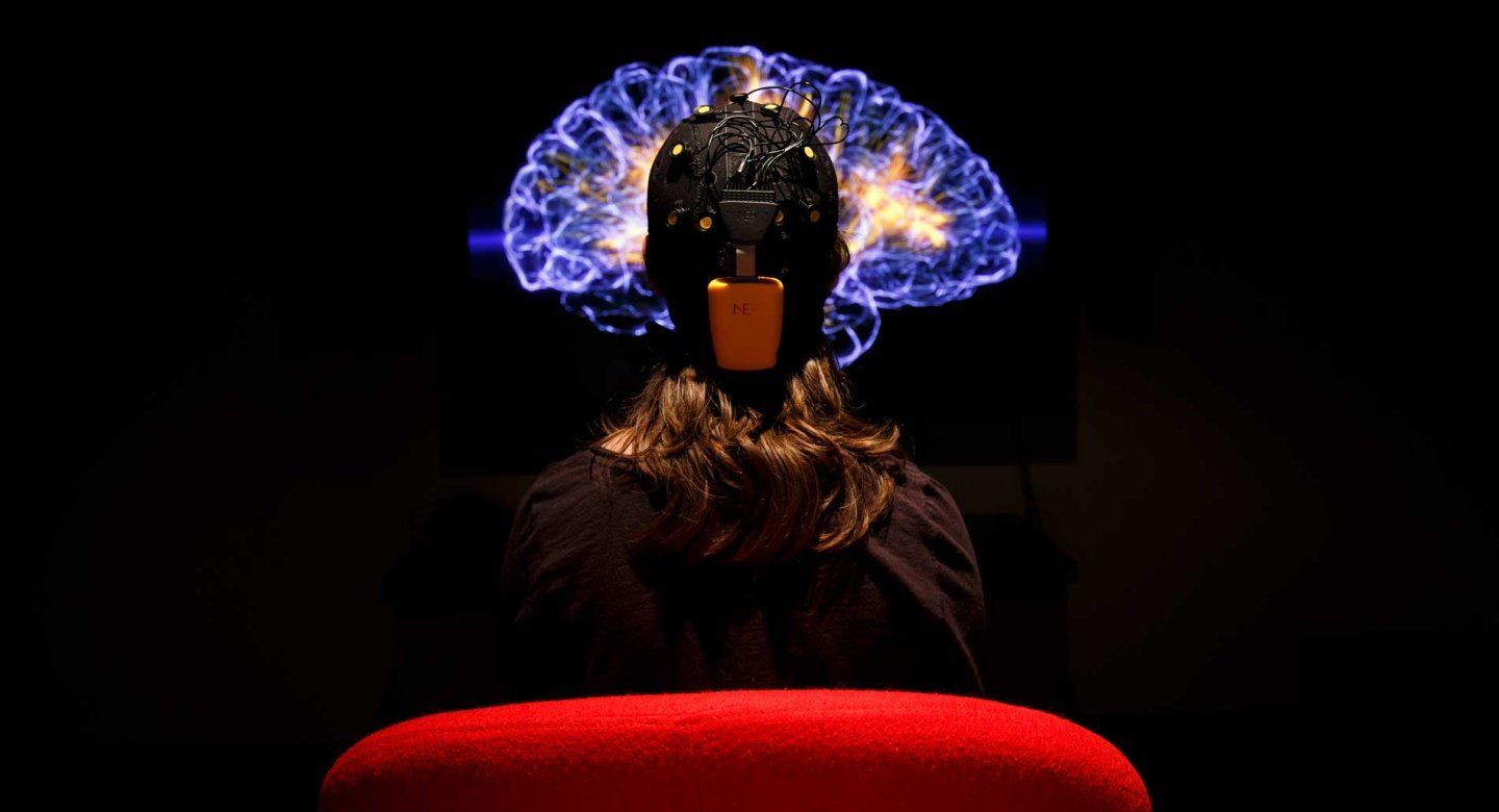 A person seen from behind sitting in a red chair and wearing a black EEG head-cap with numerous yellow sensors and wires. They are looking at a large screen in a dark room that displays a glowing, translucent blue and orange 3D visualization of a human brain.