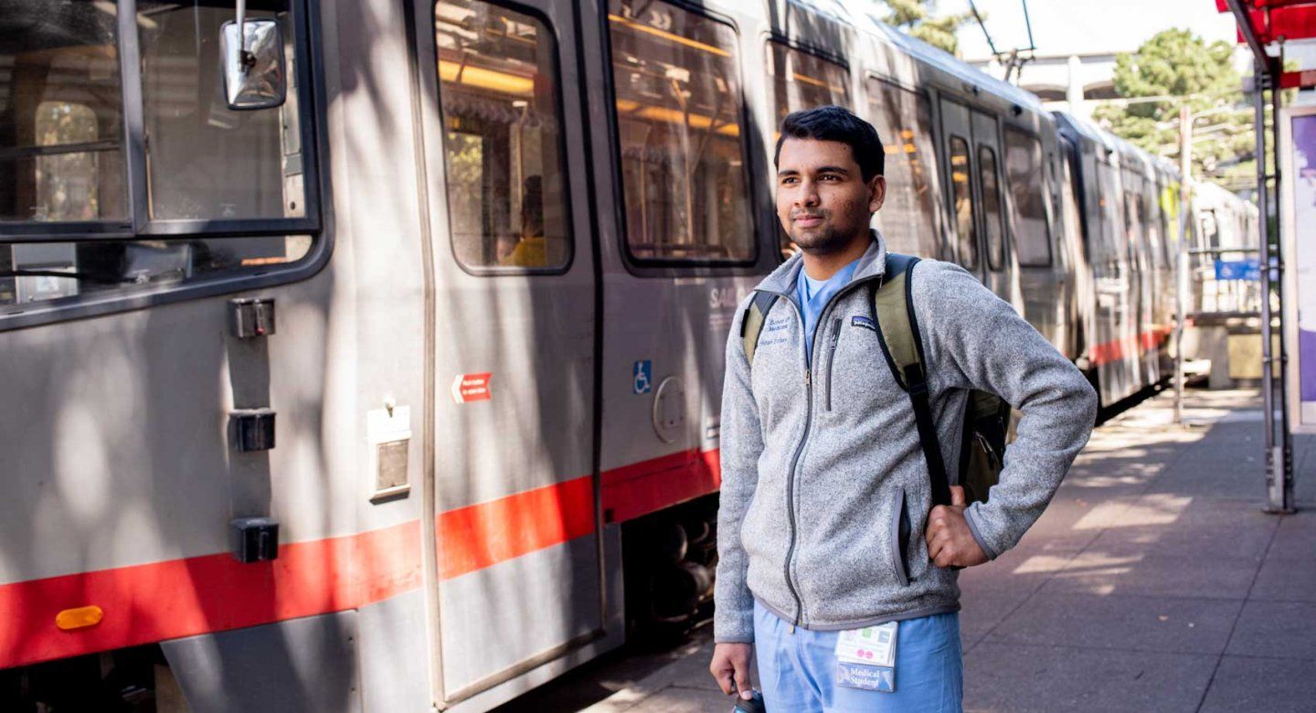 A medical student stands in front of a San Francisco MUNI rail train.