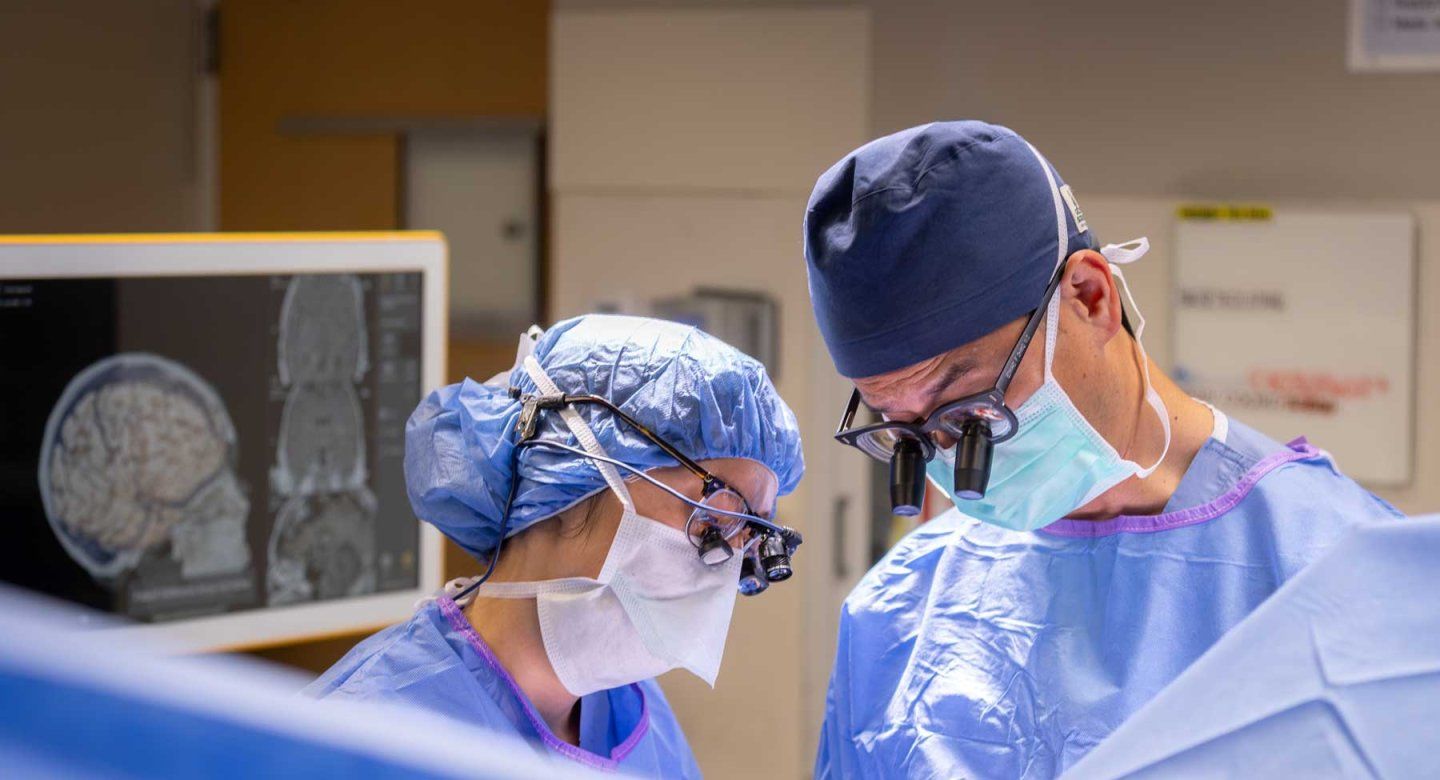 Two neurosurgeons perform surgery with a screen behind them showing a scan of a brain.