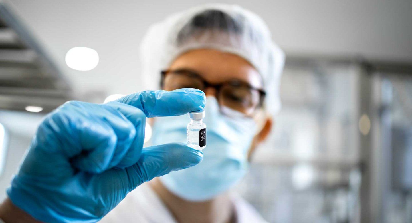 Scientist wearing protective gloves, a mask, and a hair covering examines a small vial in a laboratory.