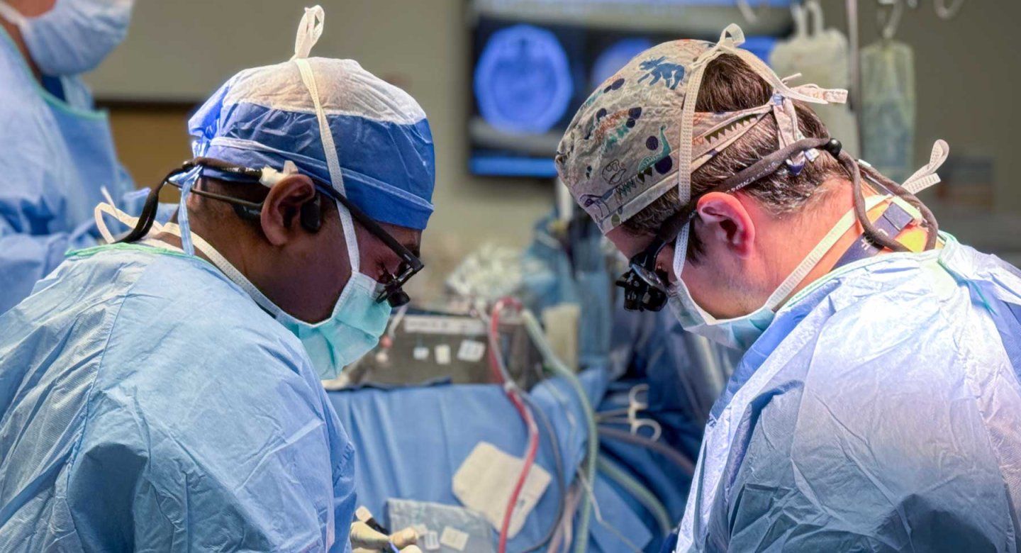 Two neurosurgeons perform surgery with a screen behind them showing a scan of a brain.