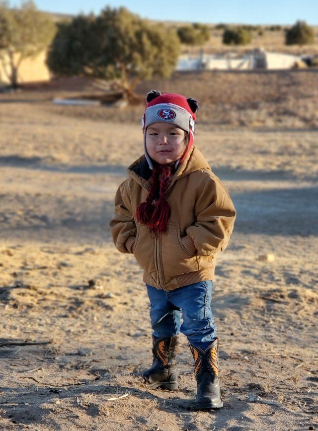 A young boy of Navajo descent smiles as he stands outdoors with his hands in his jacket, wearing a San Francisco giants beanie and cowboy boots.