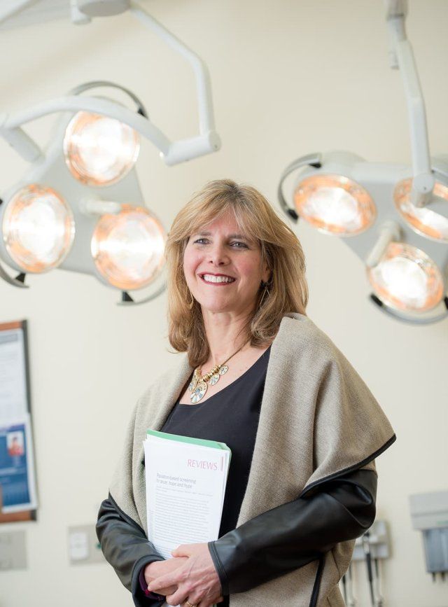 Laura Esserman stands in the middle of a medical examination room. Two sets of mounted surgical lights are in the background.