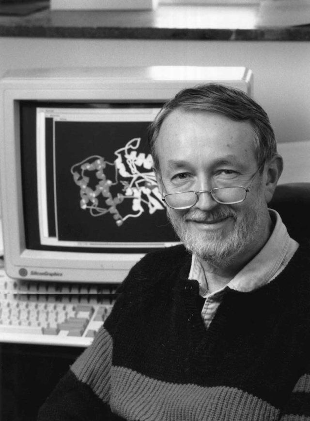 A greyscale photo of Henry Bourne. Behind him is a computer with a 3D molecular model on the screen. 