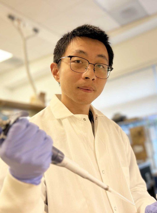 Cheng Wang, PhD, wears a lab coat and purple latex gloves as he handles scientific equipment inside a lab at UCSF.