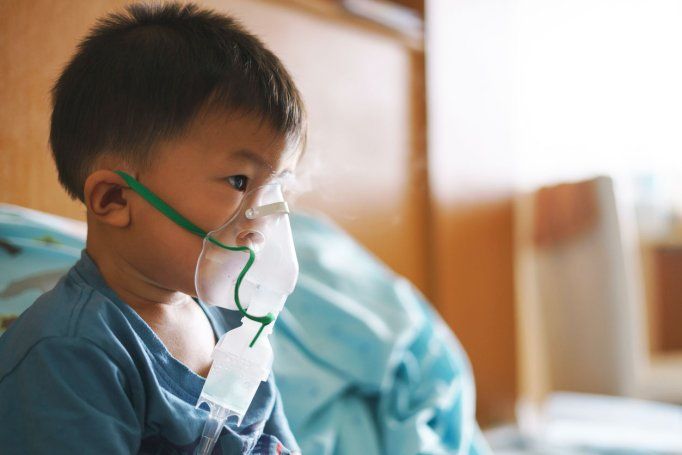 A young Asian toddler sits in a hospital using an inhaler containing medicine to stop coughing from disease like flu or RSV