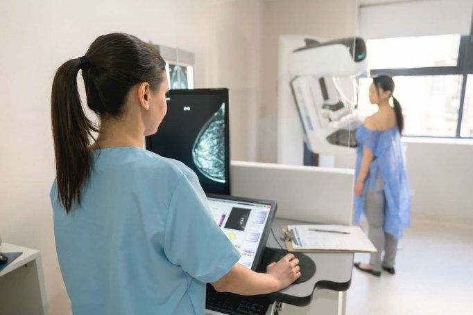 A technician takes a mammogram. The woman getting the mammogram stands at the far end of the room. The scan can be seen on the computer screen.