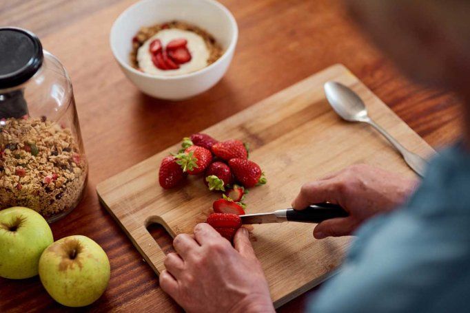 A person chops strawberries on a counter. A bowl with oatmeal, granola, and fruit is in front of them.
