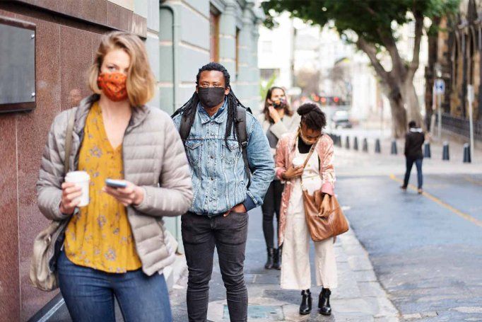 A diverse group of people wearing protective face masks practicing social distancing while waiting in a line on a city sidewalk.