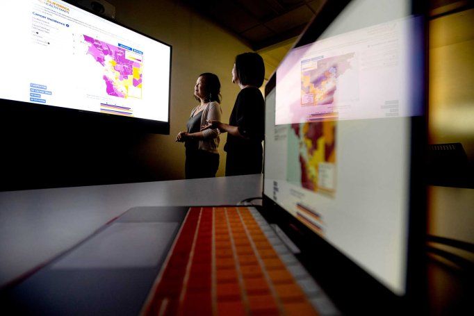 Scarlett Gomez and Debby Oh examine a screen with a map of the California Bay Area that displays cancer data. The room is dark, and in the foreground is a laptop, of which the screen reflects the researchers and the projection screen.