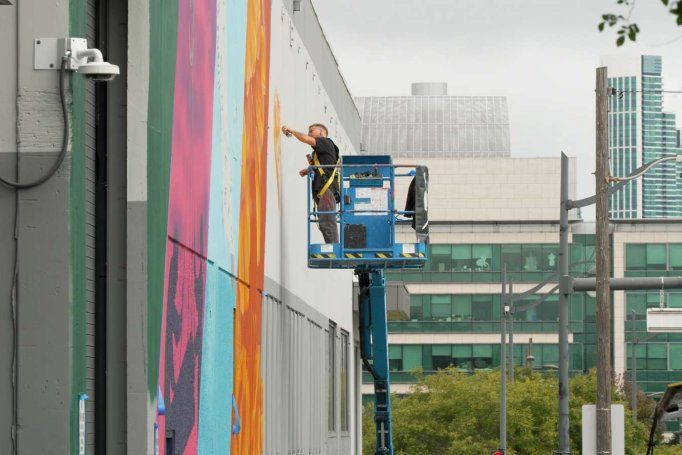 Muralist paints multi-color art on building with city skyline in background.