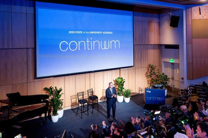 Chancellor Hawgood stands in front of audience and camera with the words "continuum" on the projector screen.