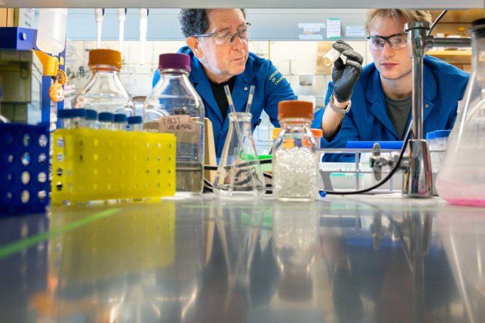 Kevan Shokat and graduate student Hunter Carrell wear blue lab coats and protective goggles as they work at a lab bench.
