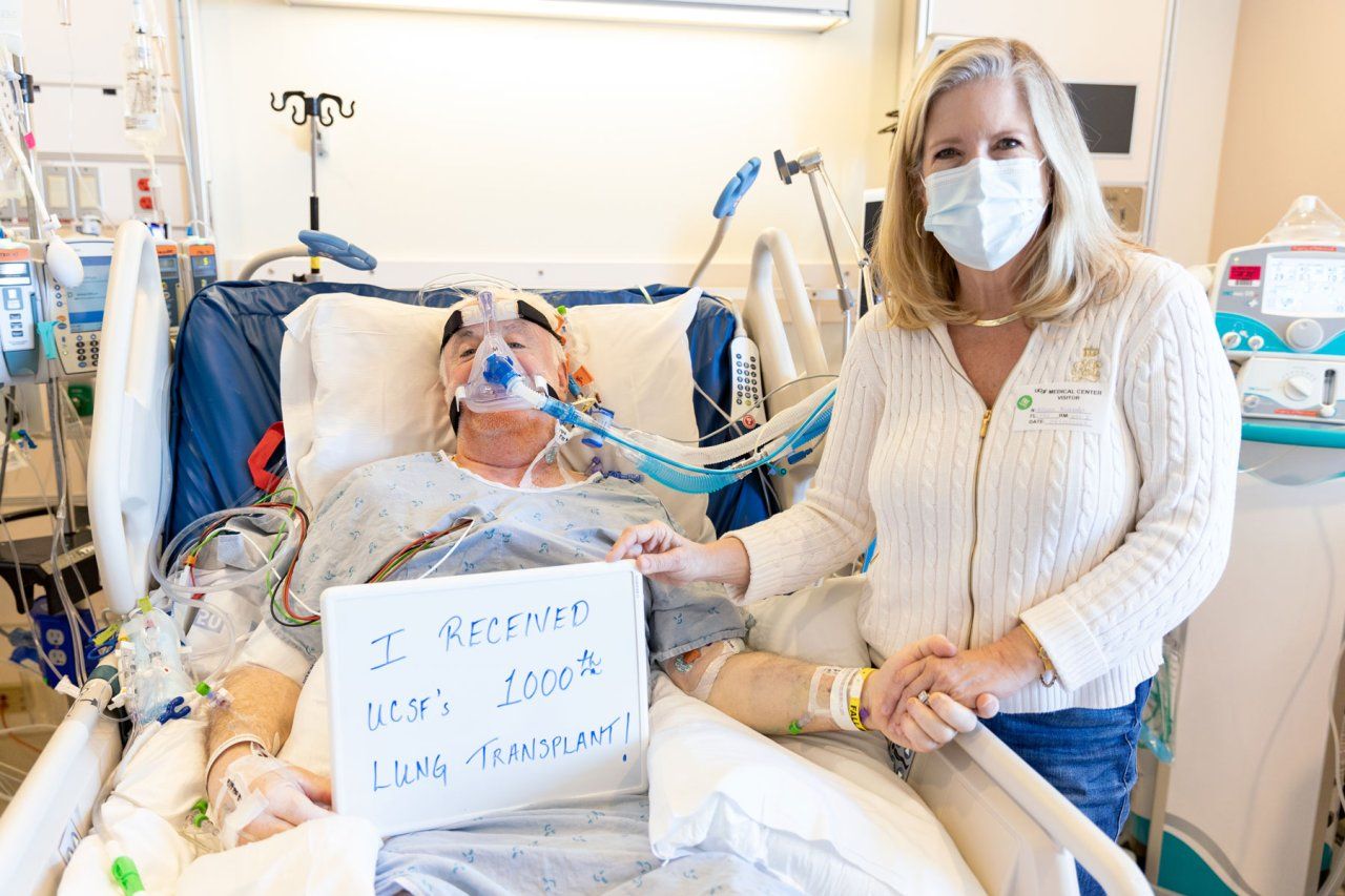 Patient Patrick lies in a hospital bed after his lung transplant surgery, with his wife Allison standing beside him. Patrick holds a sign that reads "I received UCSF's 1000th lung transplant."