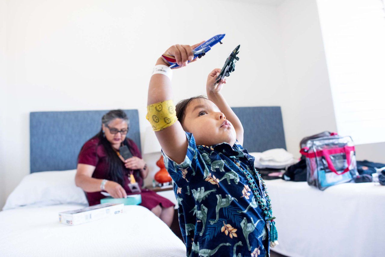 HT holds two toy airplanes aloft while his grandmother sits on a bed in the background